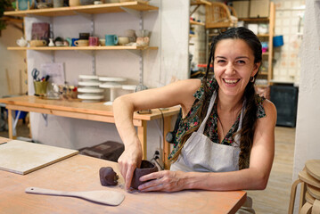 Smiling woman in studio working raw ceramic and clay to make pot. Pottery workshop with natural...