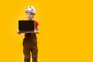 Serious schoolboy in work clothes and protective helmet holds laptop with empy screen. Copy space, mock up. Child as architect