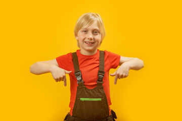 Portrait of blond boy in red T-shirt and jumpsuit showing his index fingers down and winking at the camera. Yellow background