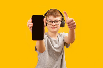 School boy in headphones and glasses holds smartphone with blank screen and shows thumbs up. Apps for listening music or learning. Copy space