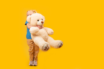 Little blond girl hugs large toy bear. Studio portrait of child with toy on bright yellow...