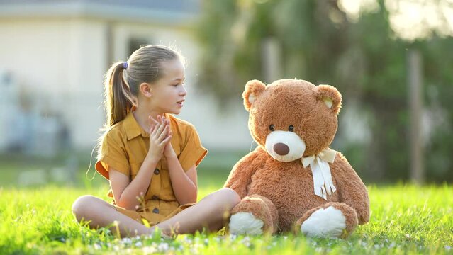 Pretty teenager girl passionately taking with her teddy bear friend outdoors on green grass lawn. Concept of friendship values