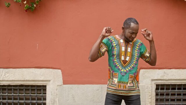 Multicultural man dancing strangely against isolated red wall background. Male in traditional African dress having fun. Mature man in ornamented shirt smiles and dances cheerfully with hands.
