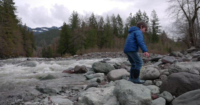Young boy exploring river bank at base of mountain on chilly autumn day - wide shot