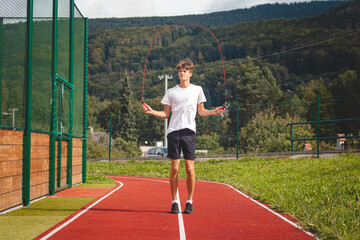 Obraz premium Brown-haired boy with an athletic figure wearing a white T-shirt and black shorts is jumping rope on an athletic oval. Training to improve jumping, coordination and endurance strength