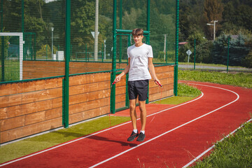 Fototapeta premium Brown-haired boy with an athletic figure wearing a white T-shirt and black shorts is jumping rope on an athletic oval. Training to improve jumping, coordination and endurance strength