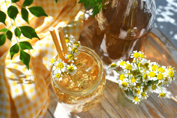 Chamomile iced tea in a mason jar with pitcher of tea and chamomile flowers on tray.