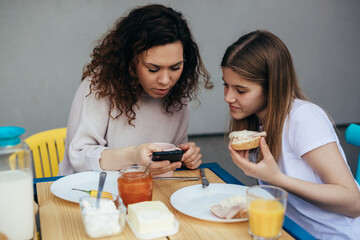 A woman and a teenage girl have sandwiches for breakfast and look at mobile phone.