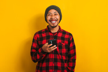 Asian man in a plaid shirt and beanie hat holding a mobile phone isolated on yellow background.