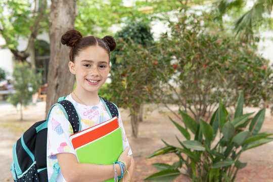 Portrait of a smiling red haired girl with a backpack and holding notebooks in the school playground looking at camera, back to school concept.