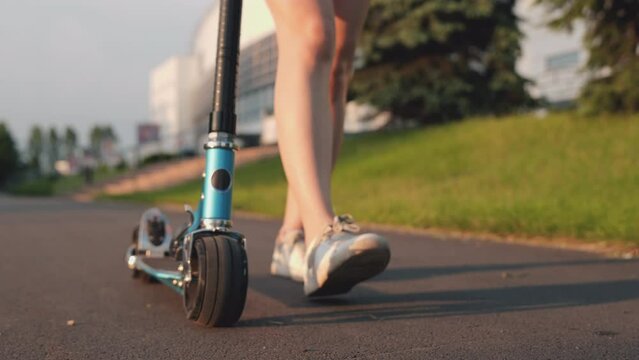 Close-up foot or leg of young woman walking on sidewalk with blue scooter. Girl walking in park on sunset. Teenager spends time alone at sunlight. Concept of college life, skating, summertime.