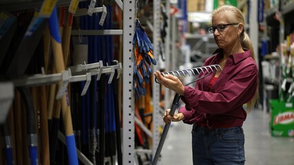 Attractive happy mature woman looking at rake garden tool and inspecting it closely in a large hardware store.