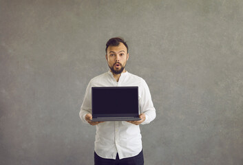 A young man stands with his mouth wide open, surprised, holding a laptop in his hands with a black screen. A handsome man in a white shirt on a black background holds a laptop.