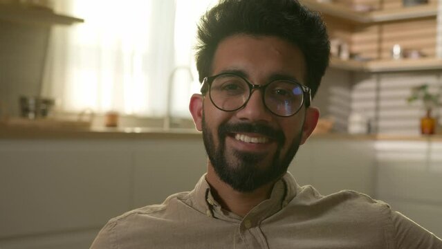 Happy smiling bearded man middle eastern Arabian millennial guy Indian male homeowner sit in domestic kitchen sun in window portrait in sunray home renter own estate apartment looking at camera smile