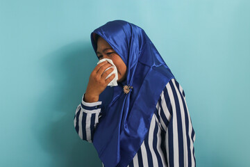 unhealthy middle-aged Asian woman, wearing a blue hijab and a striped shirt, is seen blowing her runny nose with a tissue while sneezing due to the flu. Healthcare and medical concept