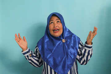 An annoyed middle-aged Asian woman, wearing a blue hijab and a striped shirt, raises her hand and...