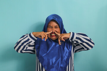 A middle-aged Asian woman, in a blue hijab, holds her breath and pinches her nose with her fingers, reacting to a stinky and disgusting, intolerable bad smell, while standing against a blue background - obrazy, fototapety, plakaty
