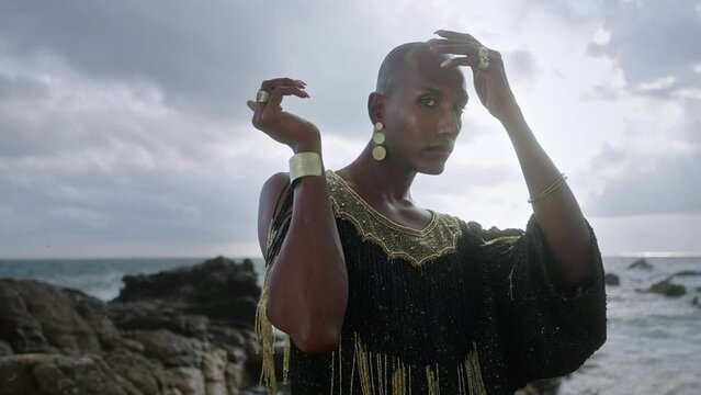 Queer black person in luxury dress, handmade jewelry posing at rocky ocean beach. Closeup slow motion. Elegant biethnic fashion gay model in posh gown, brass accessories portrait. Hand fly dance.