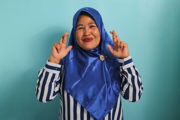 An excited middle-aged Asian woman, wearing a blue hijab, is hopeful and optimistic, crossing her fingers for good luck, making a wish, and smiling while standing against a blue background