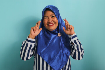 An excited middle-aged Asian woman, wearing a blue hijab, is hopeful and optimistic, crossing her fingers for good luck, making a wish, and smiling while standing against a blue background