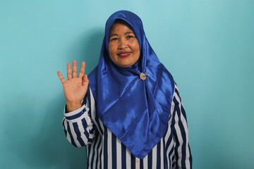 A friendly middle-aged Asian woman in a blue hijab and striped shirt is smiling, saying hi, and waving her hand to greet a person, making a hello gesture while standing against a blue background. - obrazy, fototapety, plakaty