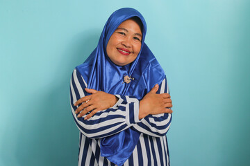 A smiling Middle-Aged Asian woman, in a blue hijab and striped shirt, embraces herself, radiating happiness, positivity, and confidence. Self-love and self-care concept, isolated on a blue background.