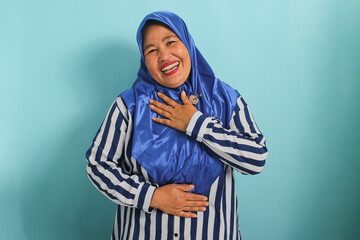 A happy middle-aged Asian woman, wearing a blue hijab and a striped shirt, holds her hands on her chest, feeling stress-free and grateful while standing against a blue background.