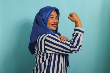 An excited middle-aged Asian woman in a blue hijab and striped shirt shows a strong gesture by lifting her arms and flexing her muscles, smiling proudly at the camera, isolated on blue background.