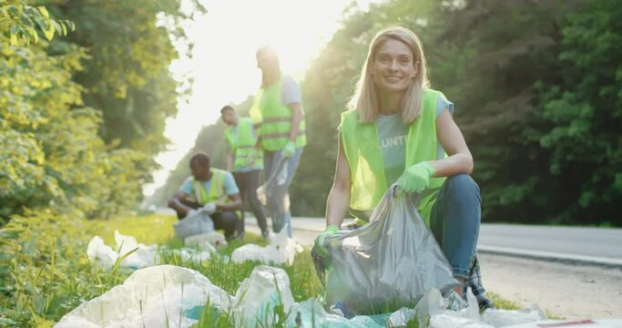 Portrait of young smiling woman with small group of volunteers on background using gloves and garbage bags for cleaning up area near road. Environment preservation and ecology concept.