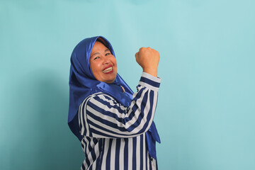 An enthusiastic middle-aged Asian woman, in a blue hijab and a striped shirt, is saying YES with a...