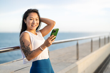 Portrait of a beautiful Asian ethnicity young sporty woman using smartphone alongside the sea shore