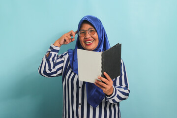 Excited Asian middle-aged woman, wearing a blue hijab, eyeglasses, and a striped shirt, holds a book and adjusts her glasses while looking directly at the camera, standing against a blue background.