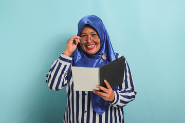 Excited Asian middle-aged woman, wearing a blue hijab, eyeglasses, and a striped shirt, holds a book and adjusts her glasses while looking directly at the camera, standing against a blue background.