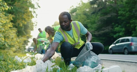 Portrait of African American man smiling and working with small group of volunteers on background. Male volunteer using gloves and garbage bags for cleaning up area near road.