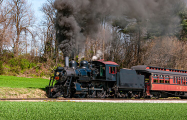 An Angled View of a Restored Steam Passenger Train Moving Slowly Blowing Lots of Black Smoke and White Steam on a Sunny Day