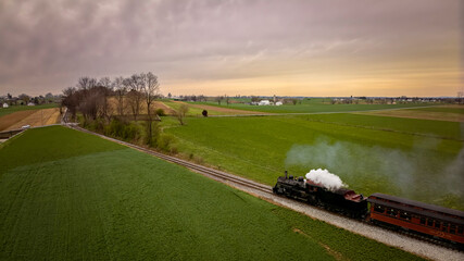 A Slightly Behind Aerial View of An Antique Passenger Steam Train Traveling on a Single Track Thru The Countryside and Farmlands, on a Spring Day