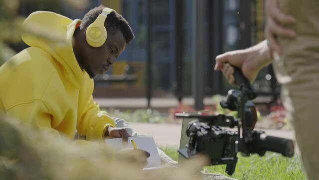 Close-up of cameraman filming dark-skinned man in headphones and yellow hoodie sitting on lawn in courtyard of residential complex with notebook in his hands. Backstage. Slow motion