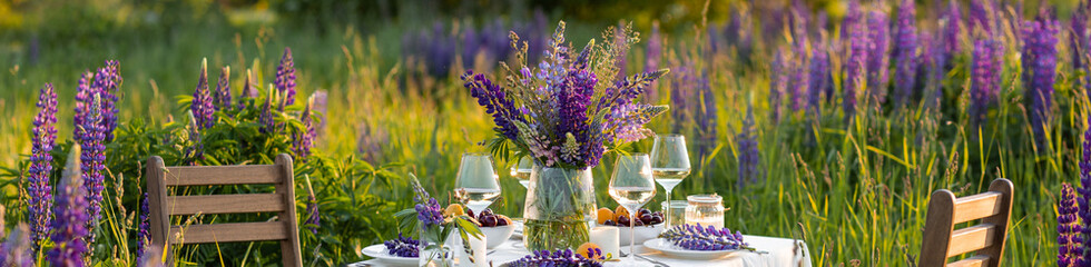 Romantic table decor for a loving couple on the blooming meadow with purple lupines. Two glasses of wine, flowers in a vase, fruits, wooden furniture and picnic basket. Sunset, golden hour. Banner