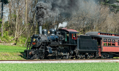An Angled View of a Restored Steam Passenger Train Moving Slowly Blowing Lots of Black Smoke and White Steam on a Sunny Day