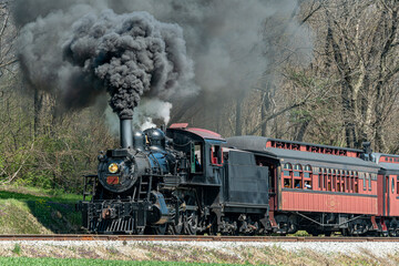 An Angled View of a Restored Steam Passenger Train Moving Slowly Blowing Lots of Black Smoke and White Steam on a Sunny Day