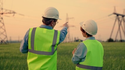 Business people, power construction. Two energy engineers work with computer tablet outdoors. Engineers in helmet, on field with electric towers, teamwork. Electrical with high voltage pylon, sunset.