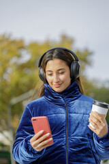 Latin girl with headphones and a disposable cup of coffee looking at her mobile phone.