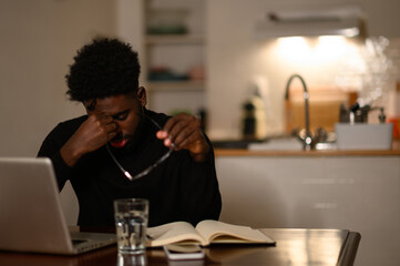 A tired multicultural hipster is sitting at home late at night and having a headache from hard work on a laptop.