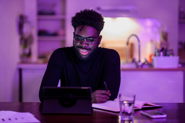 A cheerful interracial self-employed man is sitting at home late at night in a purple lighted room and having a conference call on a tablet.