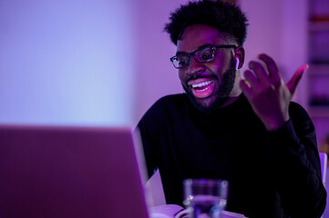A young trendy multicultural start-up businessman is sitting at home at night and having a video call with clients on a laptop.