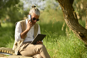 male business person drinking coffee talking on phone, doing analysis with tablet by the river in the park