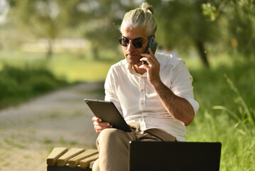 male business person drinking coffee talking on phone, doing analysis with tablet by the river in the park