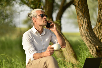 male business person drinking coffee talking on phone, doing analysis with tablet by the river in the park