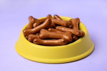Yellow bowl with bone shaped dog cookies on purple background, closeup