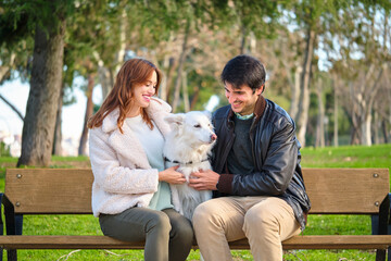 Fototapeta premium Young caucasian couple petting their dog sitting on a bench in a park. Family.
